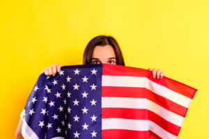 young scared latin woman covers herself with an american flag in fear of deportations, isolated on yellow background.