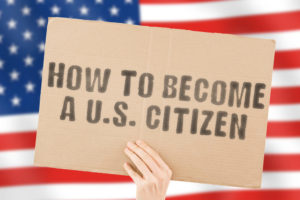 The phrase " How to become a U.S. citizen " on a banner in men's hand with blurred American flag on the background. Immigration. Nationality. Moving. Rules. Law. Legislation. Lawful. Legal