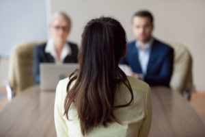 Candidate female sitting her back to camera, focus on her, close up rear view, interviewers on background.