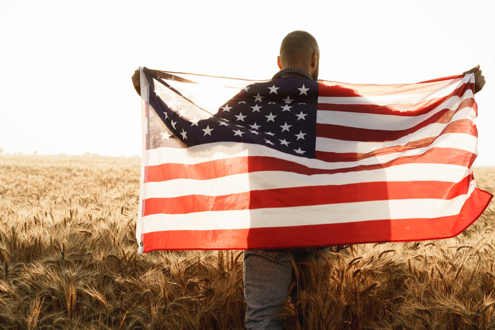 Young man holding American flag on back while standing in wheat field