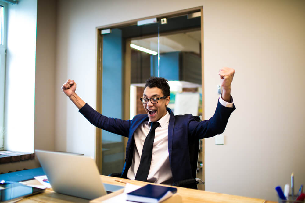 Happy smiling man winner with arms raised rejoices in its business victories watching on laptop computer, sitting in office. Cheerful entrepreneur with hands up received good news on netbook